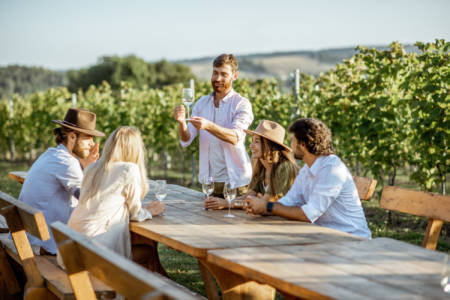 Group of a young people drinking wine and talking together while sitting at the dining table outdoors on the vineyard on a sunny evening