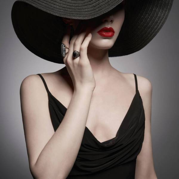 Fashion portrait of young sexy lady with beautiful black hat and evening dress. Stylish elegant woman with modern jewelry. Studio photo of pretty model on grey background.