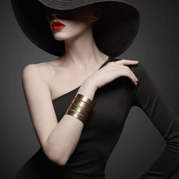 Fashion portrait of young sexy lady with beautiful black hat and evening dress. Stylish elegant woman with modern jewelry. Studio photo of pretty model on grey background.