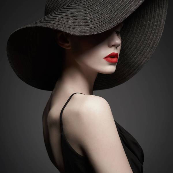 Fashion portrait of young sexy lady with beautiful black hat and evening dress. Stylish elegant woman with modern jewelry. Studio photo of pretty model on grey background.