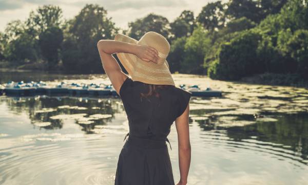 Vintage filtered shot of an elegant woman wearing a hat standing by a lake in a park at sunset