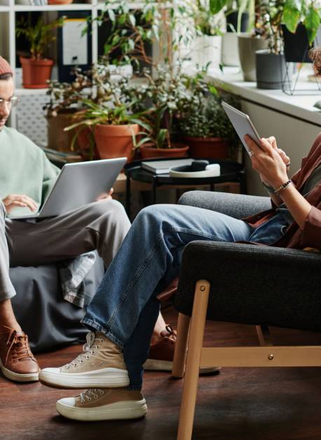 Two young serious coworkers using wireless mobile gadgets while sitting in armchairs in openspace office with variety og green plants