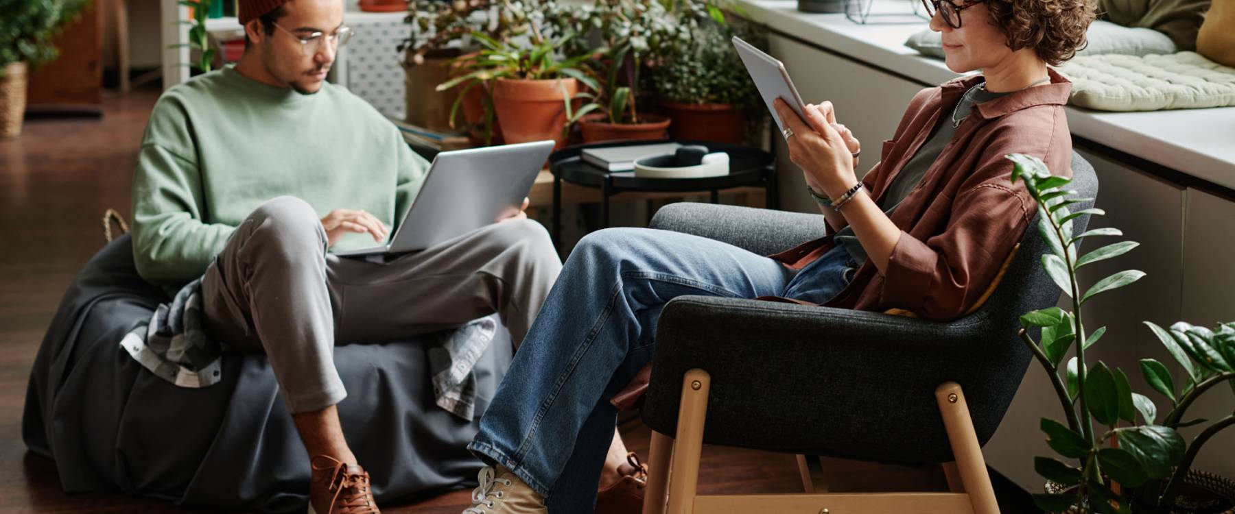 Two young serious coworkers using wireless mobile gadgets while sitting in armchairs in openspace office with variety og green plants